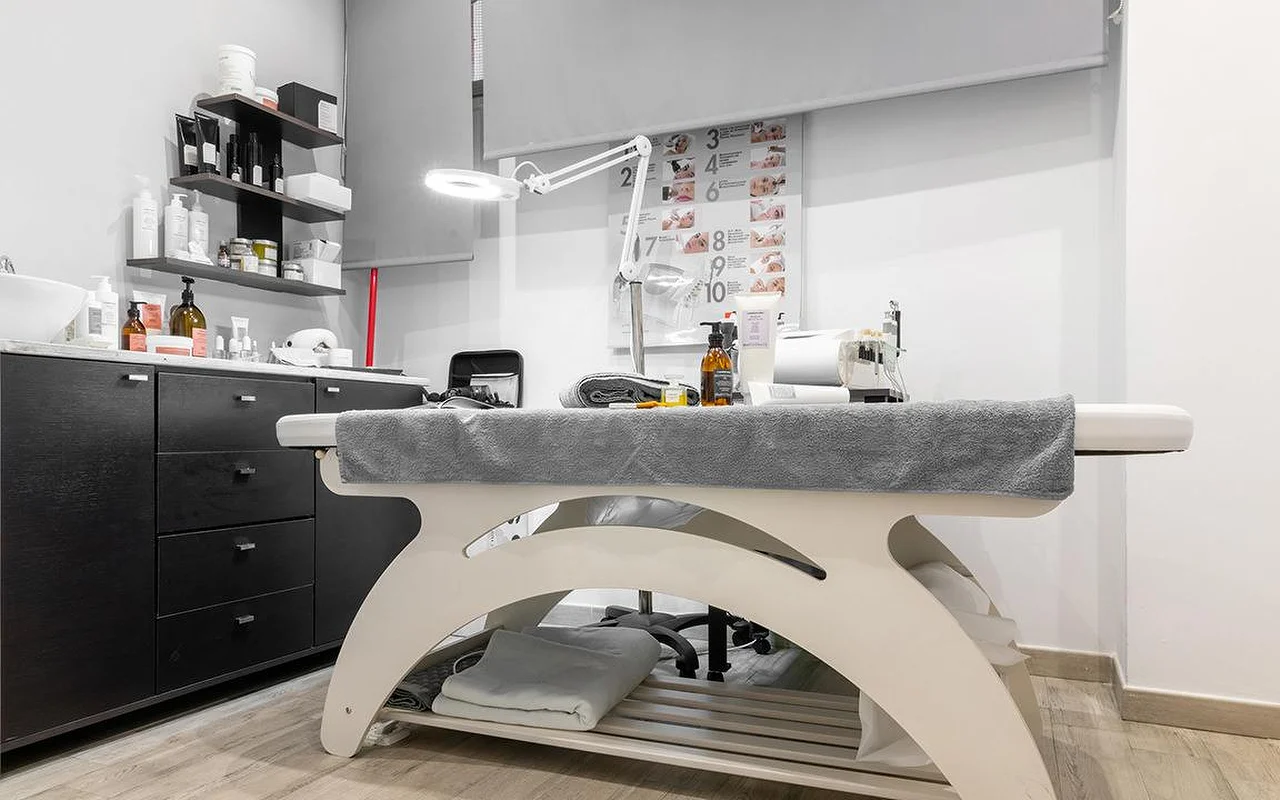 Una compatta cabina estetica da Atelier Beauty and the Lab, con un lettino color crema, una lampada d'ingrandimento regolabile, mensole scure allineate con prodotti per la pelle e, alla parete, uno schema numerato delle fasi del trattamento.