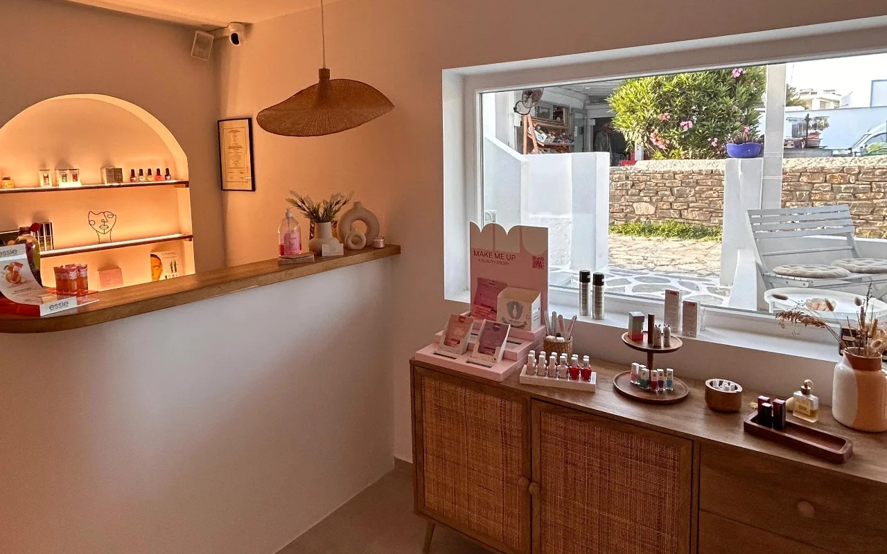 L'interno di BeautiQ Esthetics and More a Paros, con una nicchia ad arco delicatamente illuminata piena di smalti, una lampada a sospensione in rattan intrecciato e un'ampia finestra che si apre su un muro di pietra e un giardino soleggiato.