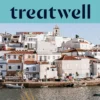 Un villaggio di pescatori dell'Algarve imbiancato a calce, con case dai tetti in cotto che salgono lungo una collina sopra uno specchio d'acqua calmo, piccole barche da pesca ormeggiate lungo la banchina, palme e il campanile di una chiesa sotto un cielo azzurro limpido.