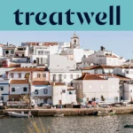 Un villaggio di pescatori dell'Algarve imbiancato a calce, con case dai tetti in cotto che salgono lungo una collina sopra uno specchio d'acqua calmo, piccole barche da pesca ormeggiate lungo la banchina, palme e il campanile di una chiesa sotto un cielo azzurro limpido.