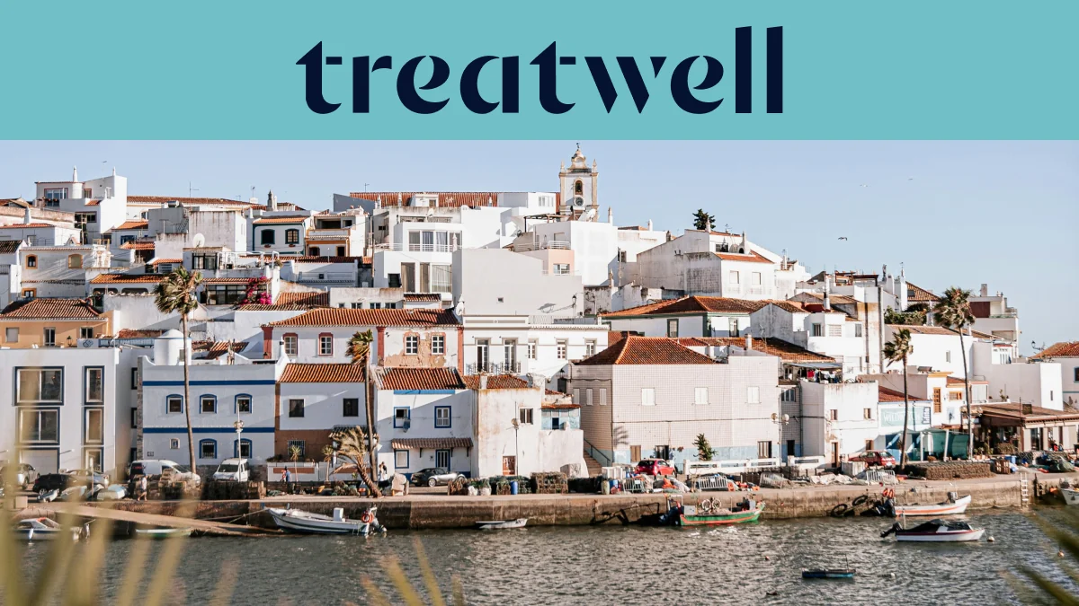Un villaggio di pescatori dell'Algarve imbiancato a calce, con case dai tetti in cotto che salgono lungo una collina sopra uno specchio d'acqua calmo, piccole barche da pesca ormeggiate lungo la banchina, palme e il campanile di una chiesa sotto un cielo azzurro limpido.