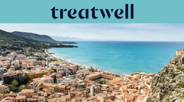 Ampia veduta su una cittadina costiera siciliana, con edifici antichi dai tetti in cotto, una mezzaluna di spiaggia sabbiosa, mare turchese cristallino e un promontorio roccioso che si erge su un lato.