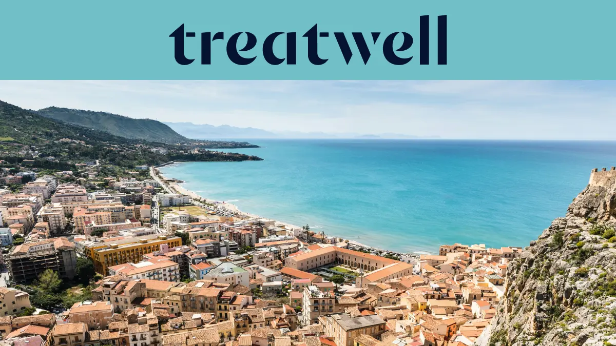 Ampia veduta su una cittadina costiera siciliana, con edifici antichi dai tetti in cotto, una mezzaluna di spiaggia sabbiosa, mare turchese cristallino e un promontorio roccioso che si erge su un lato.