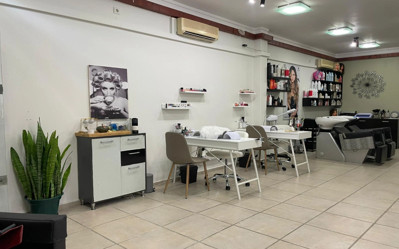 Un salone di capelli e bellezza con due tavoli da manicure bianchi e sedie color tortora, un lavatesta e mensole di prodotti lungo la parete, un'alta sansevieria in un angolo e una grande fotografia in bianco e nero di una donna con i bigodini.