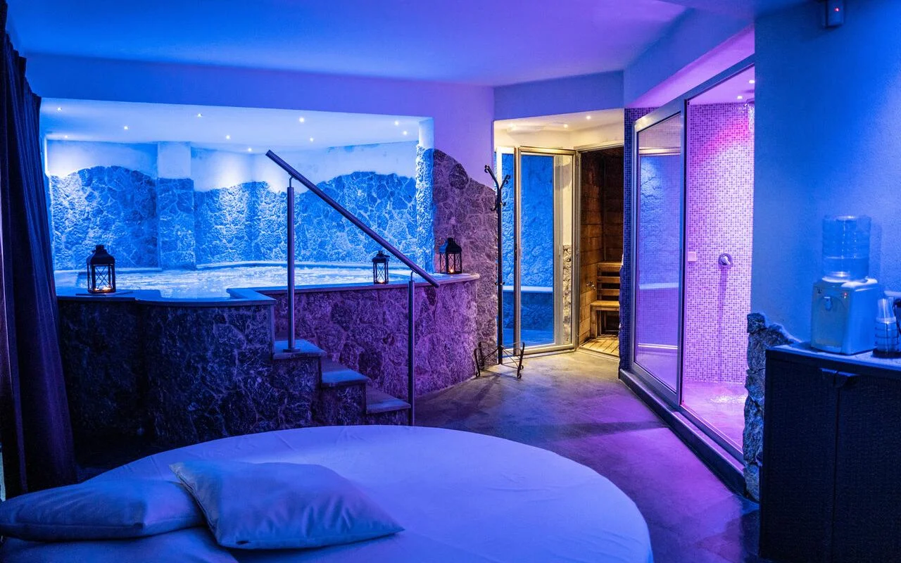 Una spa privata da Bellezza e Benessere a Taormina, illuminata di blu e viola, con una vasca idromassaggio incassata in un rivestimento in pietra, una sauna in legno e, sul fondo, un bagno turco a mosaico, e un letto rotondo in primo piano.