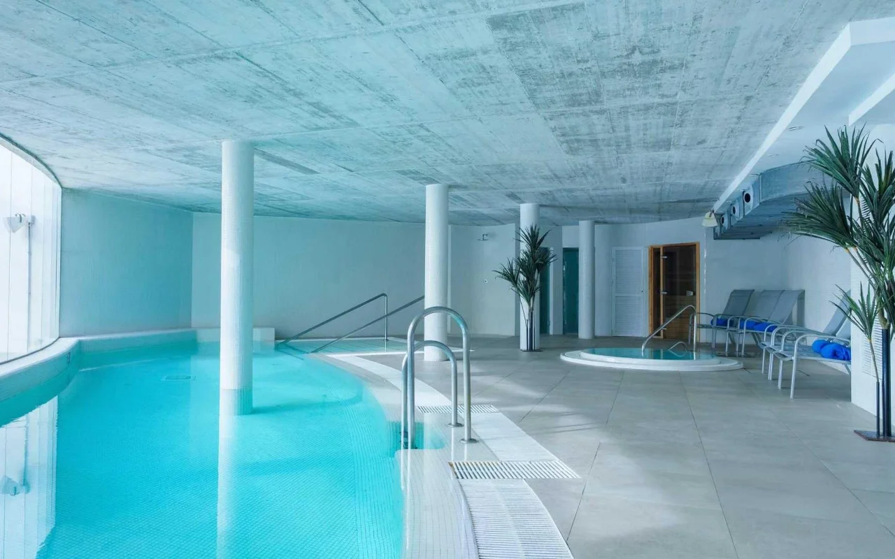La piscina interna della spa di Benalmádena Beach, con una vasca turchese curva, un soffitto in cemento a vista, una piccola jacuzzi e lettini tra alte palme in vaso.