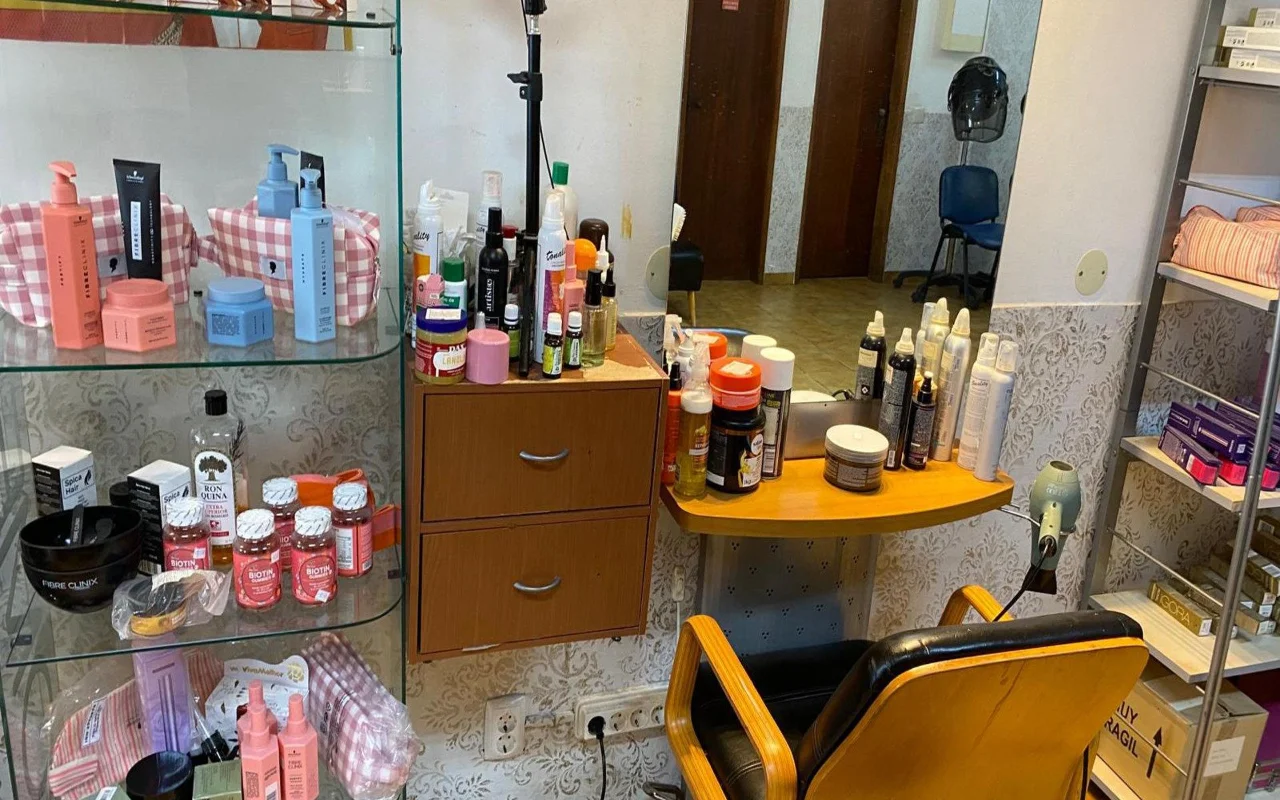 La postazione di un parrucchiere con uno specchio a parete e un piano in legno affollato di prodotti per lo styling, una vetrinetta di prodotti per la cura dei capelli e integratori accanto, e una classica poltrona da parrucchiere in legno e nero.