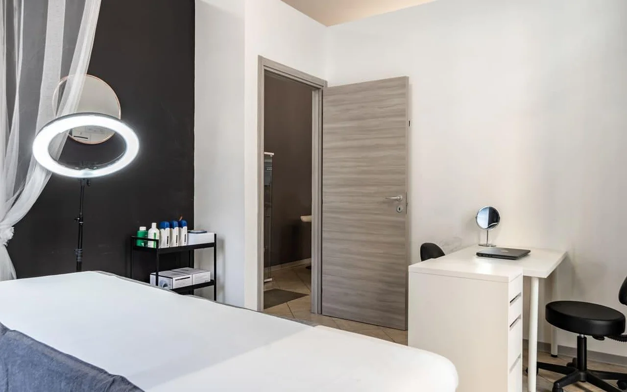 Una cabina da Coquette Wellness and Beauty, con un lettino bianco, una ring light illuminata su piantana contro una parete nera a effetto, tende leggere alla finestra e una piccola scrivania bianca con uno specchio da trucco accanto a una porta che dà su un bagno privato.
