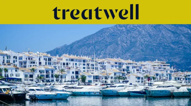 Il porto turistico di Puerto Banús, a Marbella, circondato da edifici bianchi e yacht ormeggiati sullo sfondo delle montagne, sotto uno striscione giallo Treatwell.