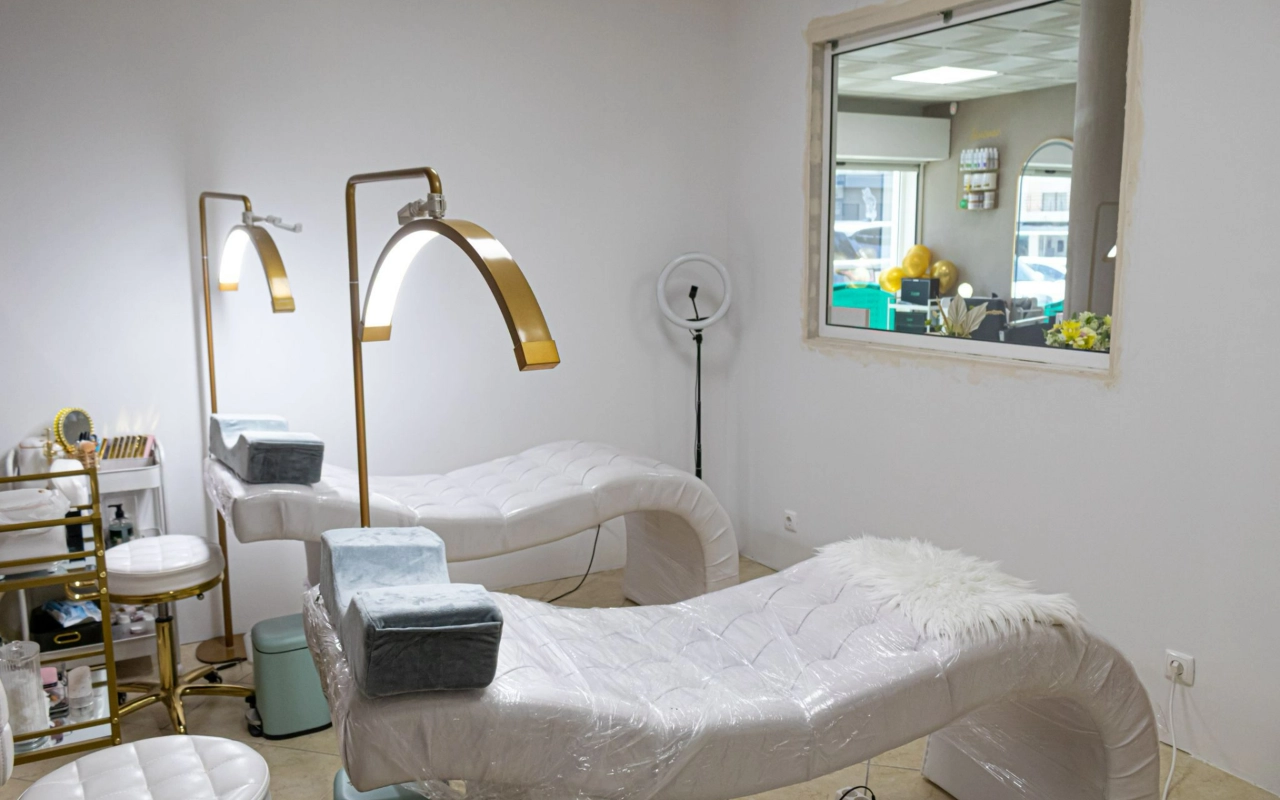 Una luminosa sala trattamenti beauty con due lettini bianchi trapuntati, alte lampade ad arco dorate e una ring light, con una finestra interna che si affaccia sulla reception.