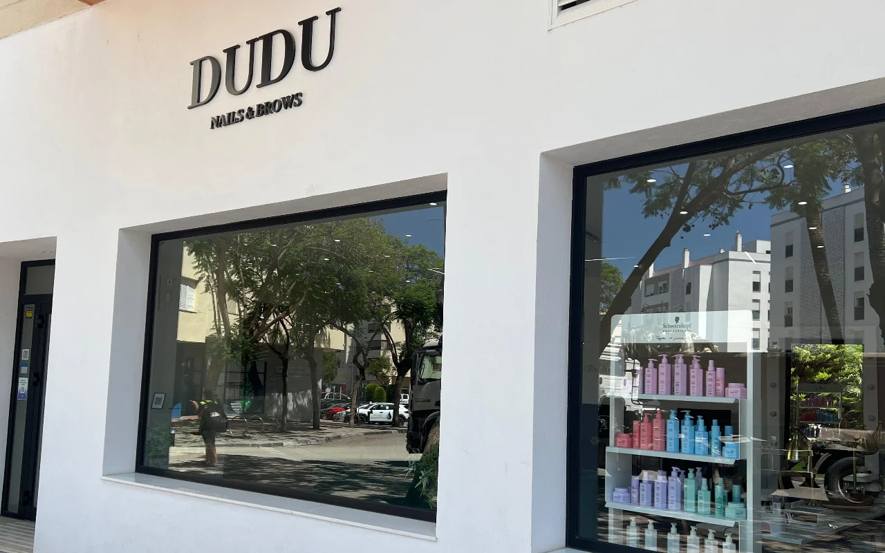La vetrina bianca di Dudu Nails and Brows a Estepona, con il nome a lettere scure sopra ampie vetrine che mostrano file di prodotti per capelli.