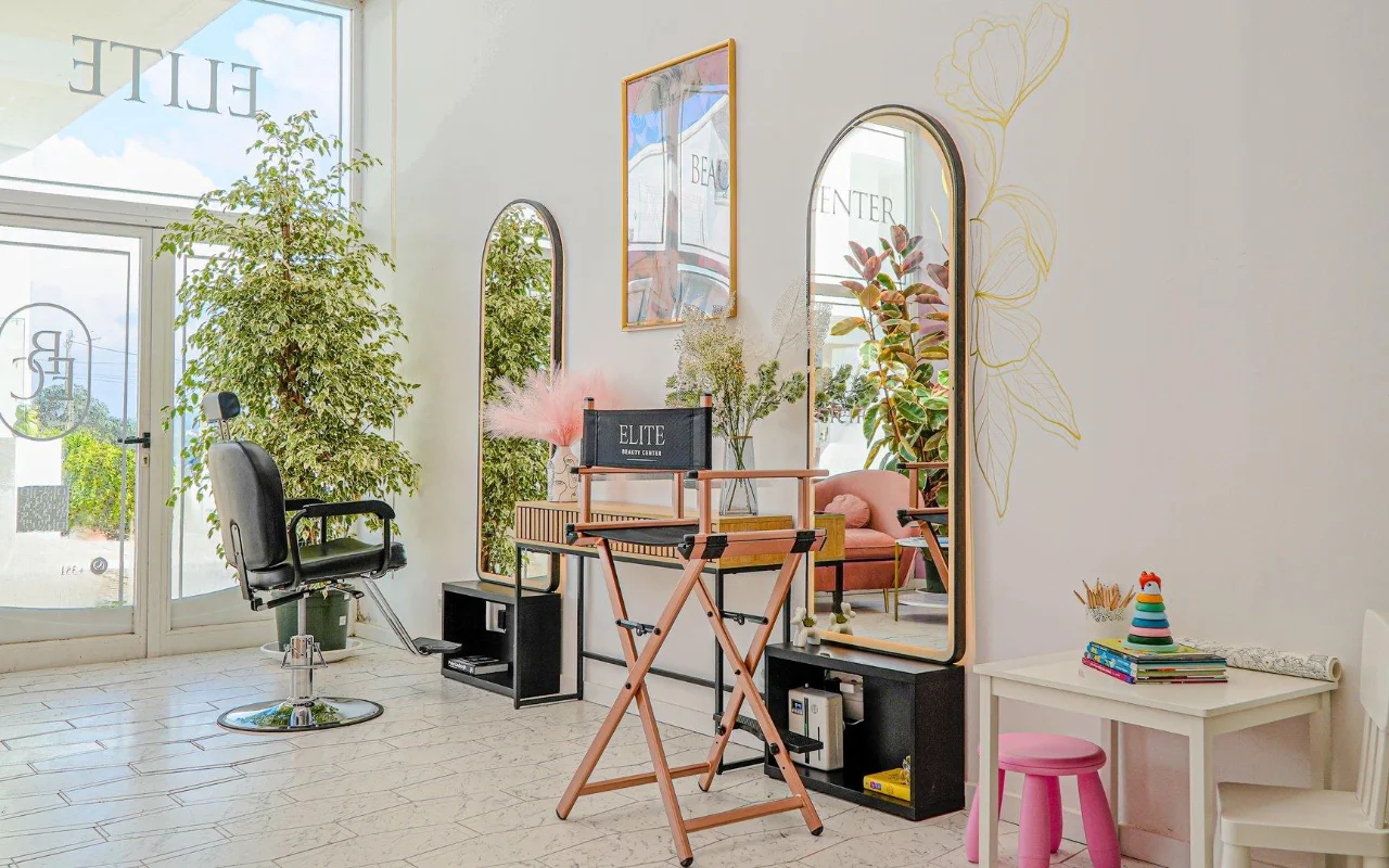 Un salone di bellezza luminoso e arioso, tutto bianco, con specchi ad arco dalle cornici dorate, una poltrona da parrucchiere nera e una sedia pieghevole da regista, alte piante verdi, accenti rosa e un piccolo angolo bimbi vicino alla finestra.