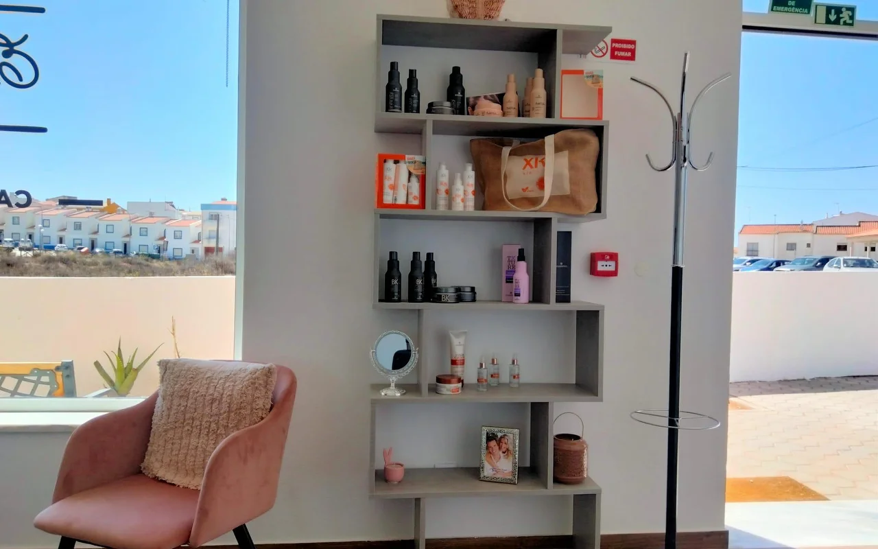 Un angolo di salone accanto a una grande finestra, con una mensola grigia a gradini piena di prodotti per capelli e una foto incorniciata, una poltrona in velluto rosa cipria con un cuscino, e la vista su basse case imbiancate a calce sotto un cielo azzurro.