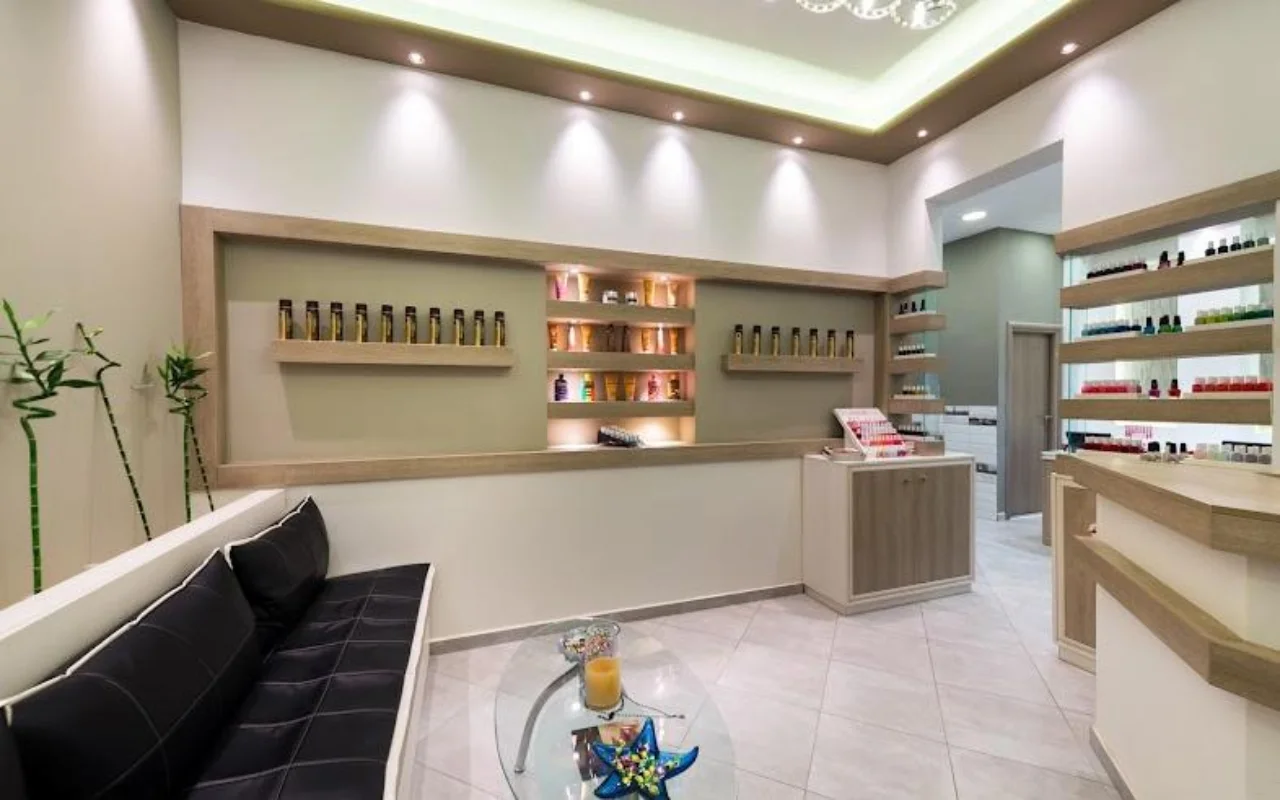 La reception dai toni crema di Fabulous Nails and More a Fira, Santorini, con una panca in pelle nera, un tavolino di vetro, mensole retroilluminate di prodotti di bellezza e una luce soffusa a soffitto.