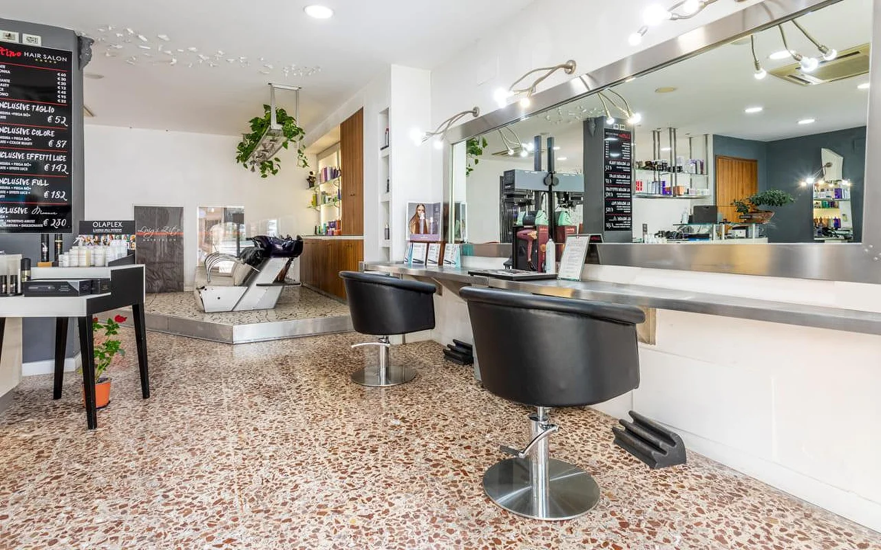 L'interno di Hair Salon Luigi Zito, con pavimento in terrazzo alla veneziana, due poltrone da styling nere davanti a un lungo specchio, listini prezzi su lavagna alla parete, un espositore di prodotti e un lavatesta sul fondo.