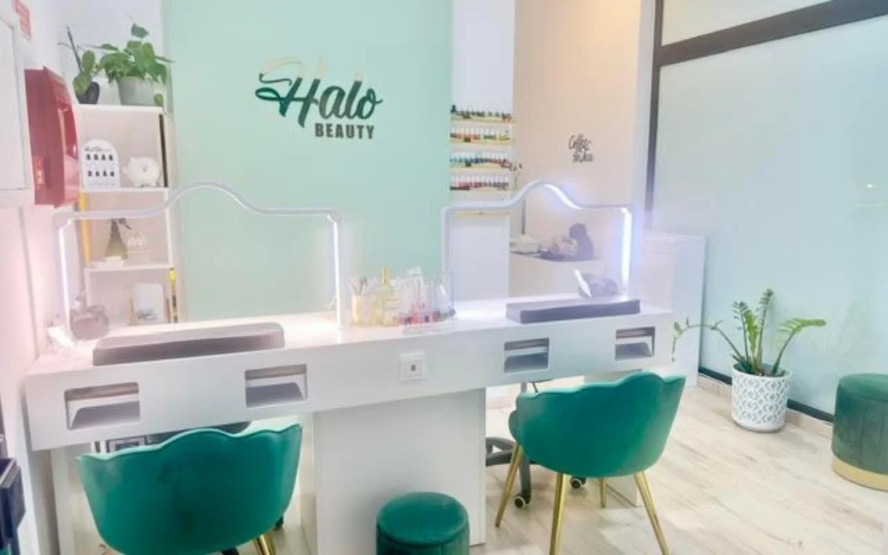 Un salone unghie con una parete d'accento verde menta che riporta il logo Halo Beauty, un doppio bancone da manicure bianco con divisori in plexiglass illuminati, due poltroncine a botte in velluto verde smeraldo, un espositore a parete di smalti e piante in vaso.