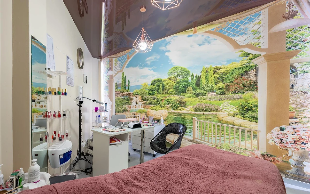 Una sala trattamenti beauty con un lettino drappeggiato di rosa e una postazione unghie bianca, davanti a un grande murale a parete di giardino e lago, con mensole in vetro rifornite di prodotti accanto.