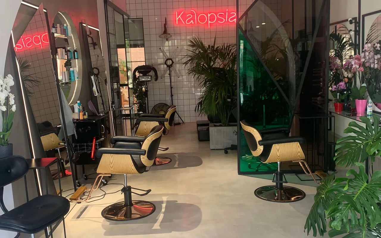 Lo spazio styling del salone Kalopsia a Ermoupoli, Syros, con poltrone in legno chiaro e nero, specchi ad arco retroilluminati, piante frondose e un'insegna al neon rosa con scritto Kalopsia.
