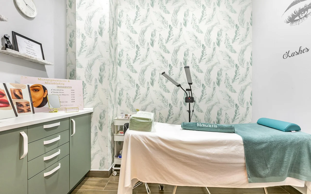 Una cabina trattamenti da Masami Belleza Natural a Fuengirola, con un lettino, una carta da parati a piume, una lampada con lente d'ingrandimento e un listino prezzi di micropigmentazione.