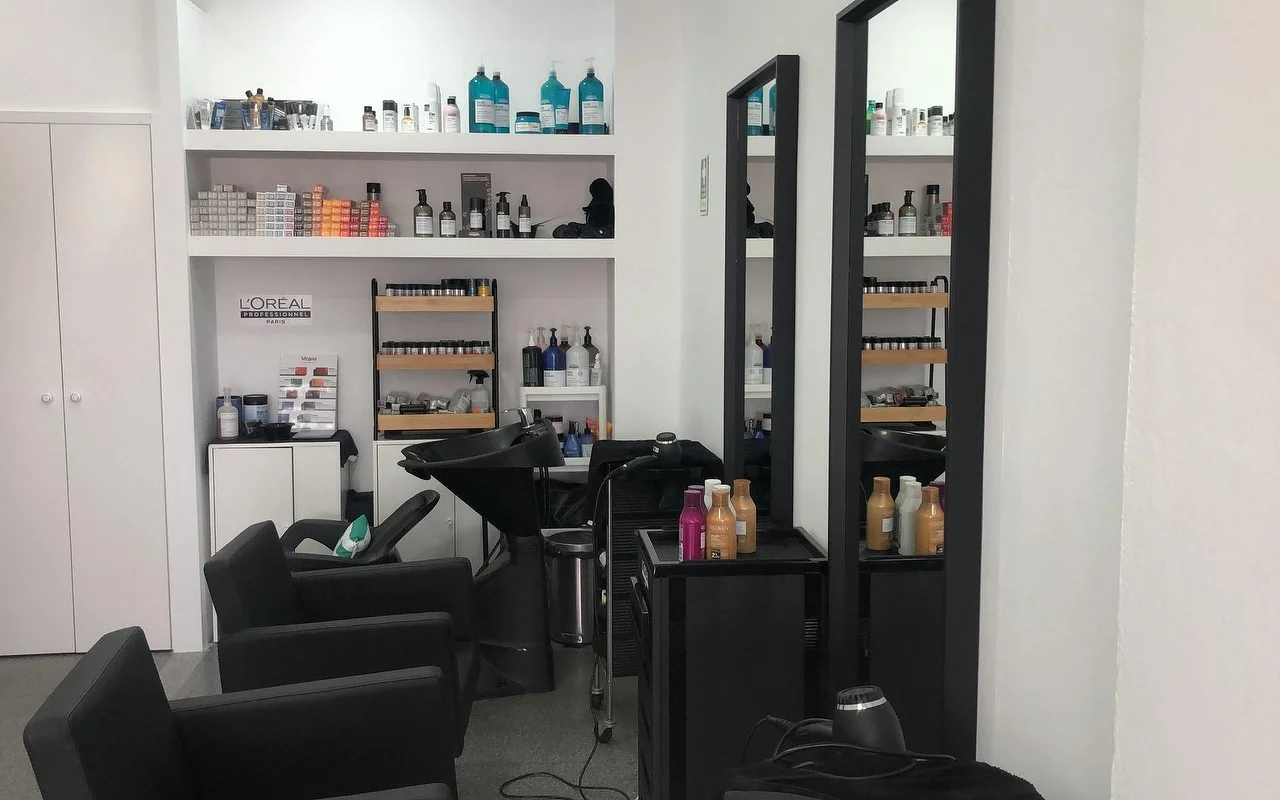 Un salone di parrucchiere con poltrone e lavatesta neri, due specchi a parete alti e stretti, e mensole rifornite di prodotti professionali di colore e cura dei capelli.