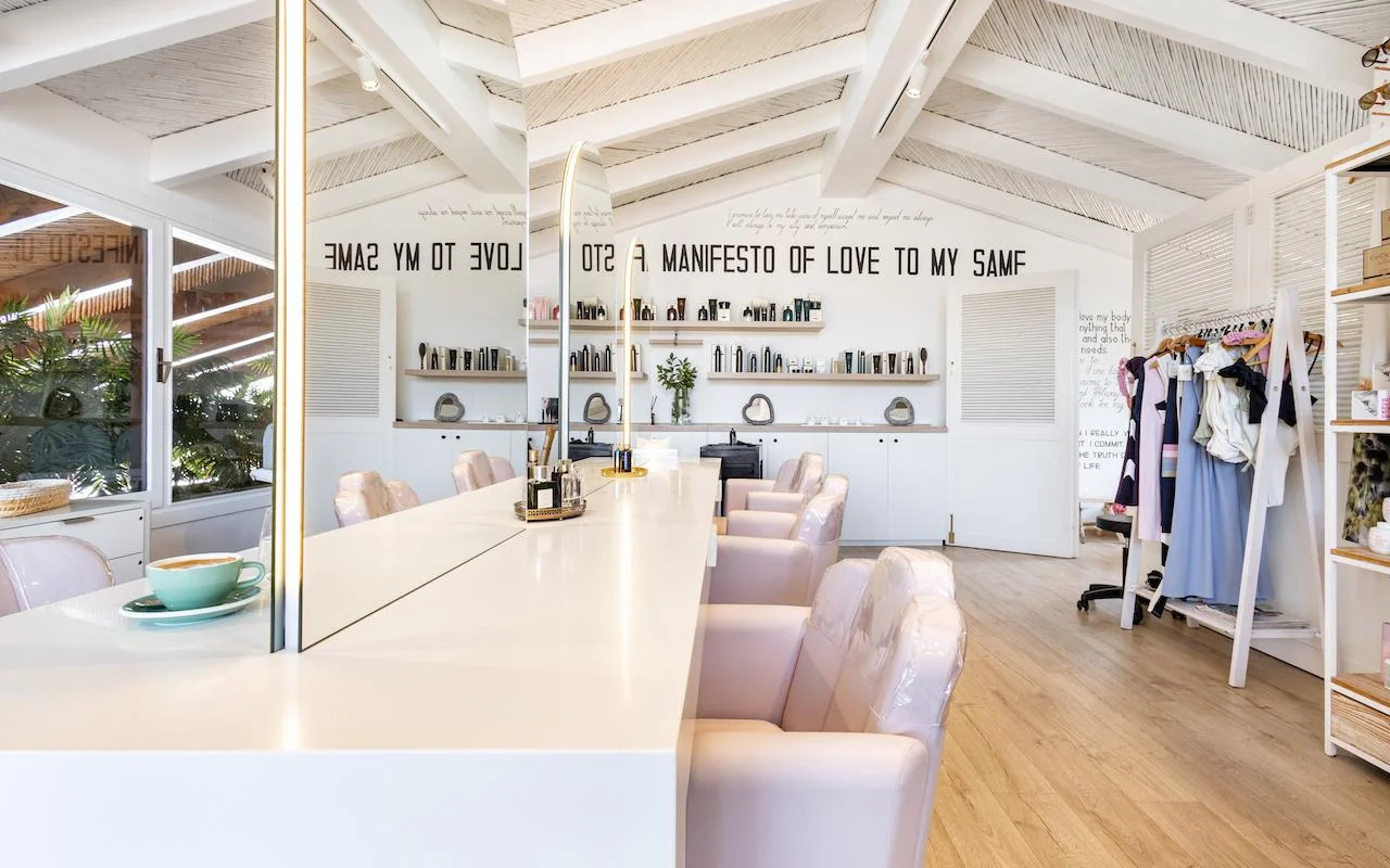 L'interno luminoso di Milk & Roses a Puente Romano, Marbella, con un bancone bianco, poltrone rosa pallido, un soffitto con travi e mensole di prodotti lungo la parete.