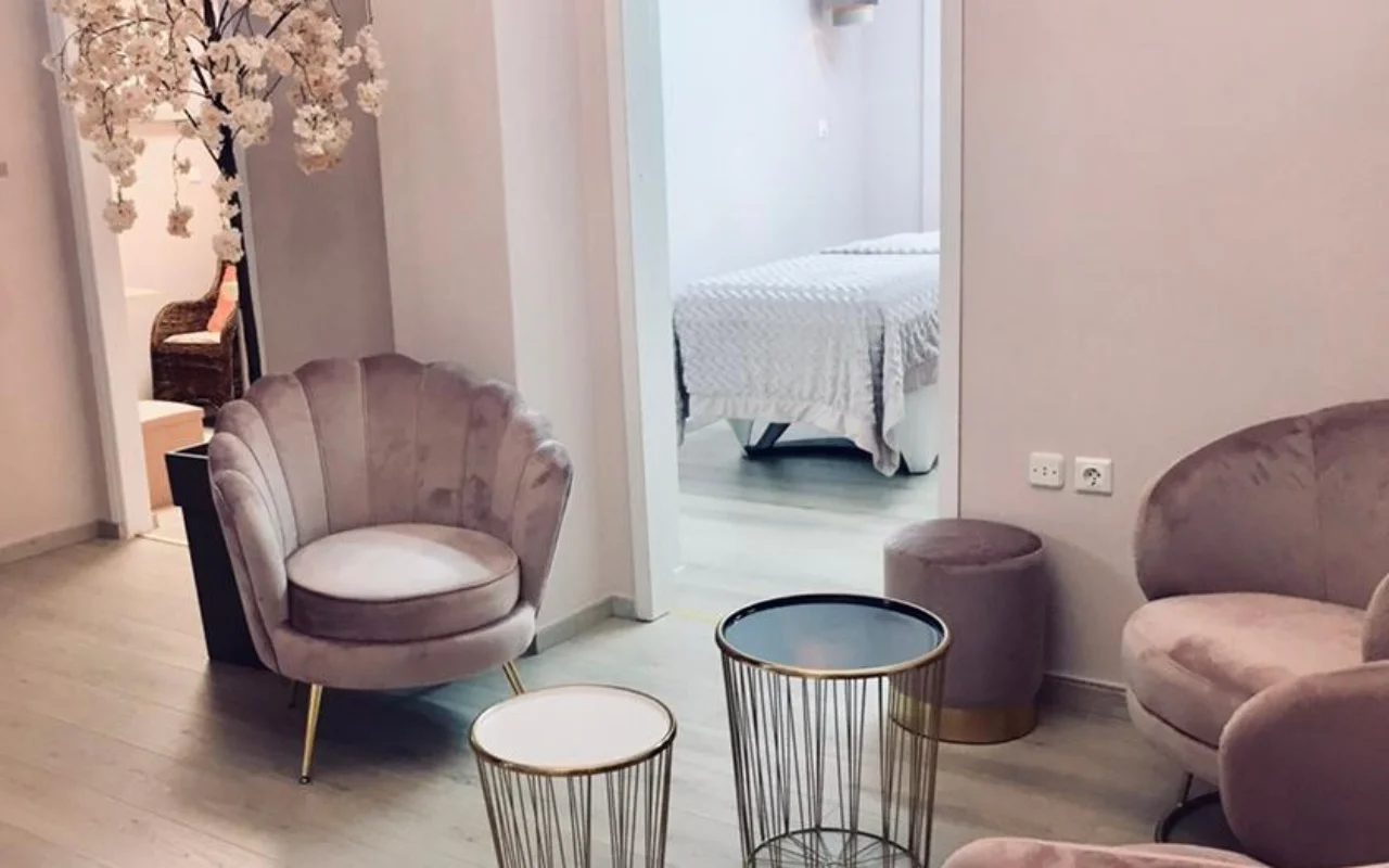 La zona d'attesa rosa cipria di Nomikou Antonia Aesthetic Lab a Syros, con una poltrona in velluto dallo schienale smerlato, sottili tavolini dorati, un ramo di ciliegio in fiore e una cabina trattamenti oltre la porta.