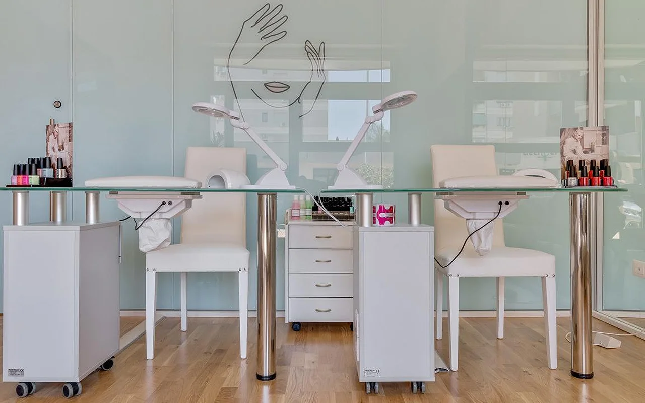 Uno studio unghie da Perfetta Urban Beauty, con due tavoli da manicure dal piano in vetro e sedie bianche, lampade da lavoro, espositori di smalti colorati e una parete in vetro verde menta con il disegno a linee di un volto e di due mani.
