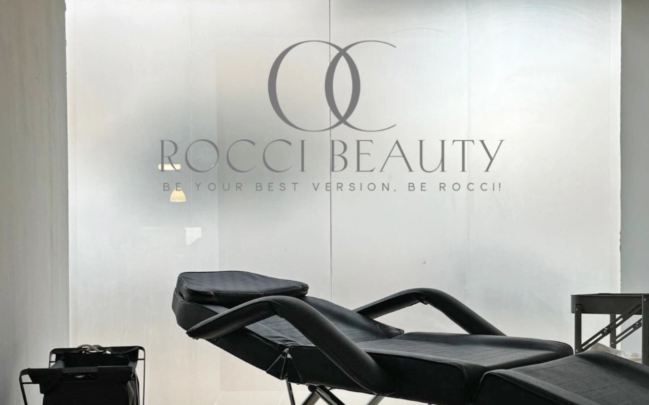 Una sala trattamenti con un lettino reclinabile nero e un piccolo carrello attrezzato, davanti a una parete in vetro smerigliato che riporta il logo e il claim di Rocci Beauty.