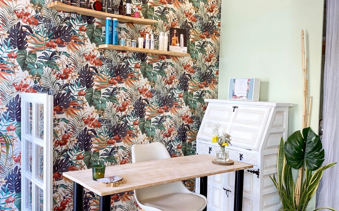 Una postazione da lavoro da Shangara Lora Organics a Marbella, con una carta da parati tropicale audace, un tavolo di legno, un antico secrétaire bianco e prodotti bio per capelli su mensole sospese.