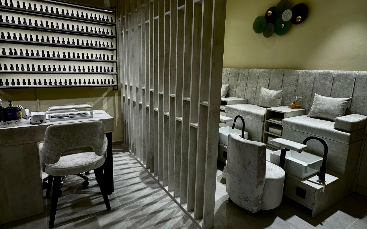 Una cabina trattamenti da Sisu Nail Club and Academy a Perissa, Santorini, con un'intera parete di smalti, poltrone per manicure e pedicure in velluto grigio e un divisorio in cemento traforato.