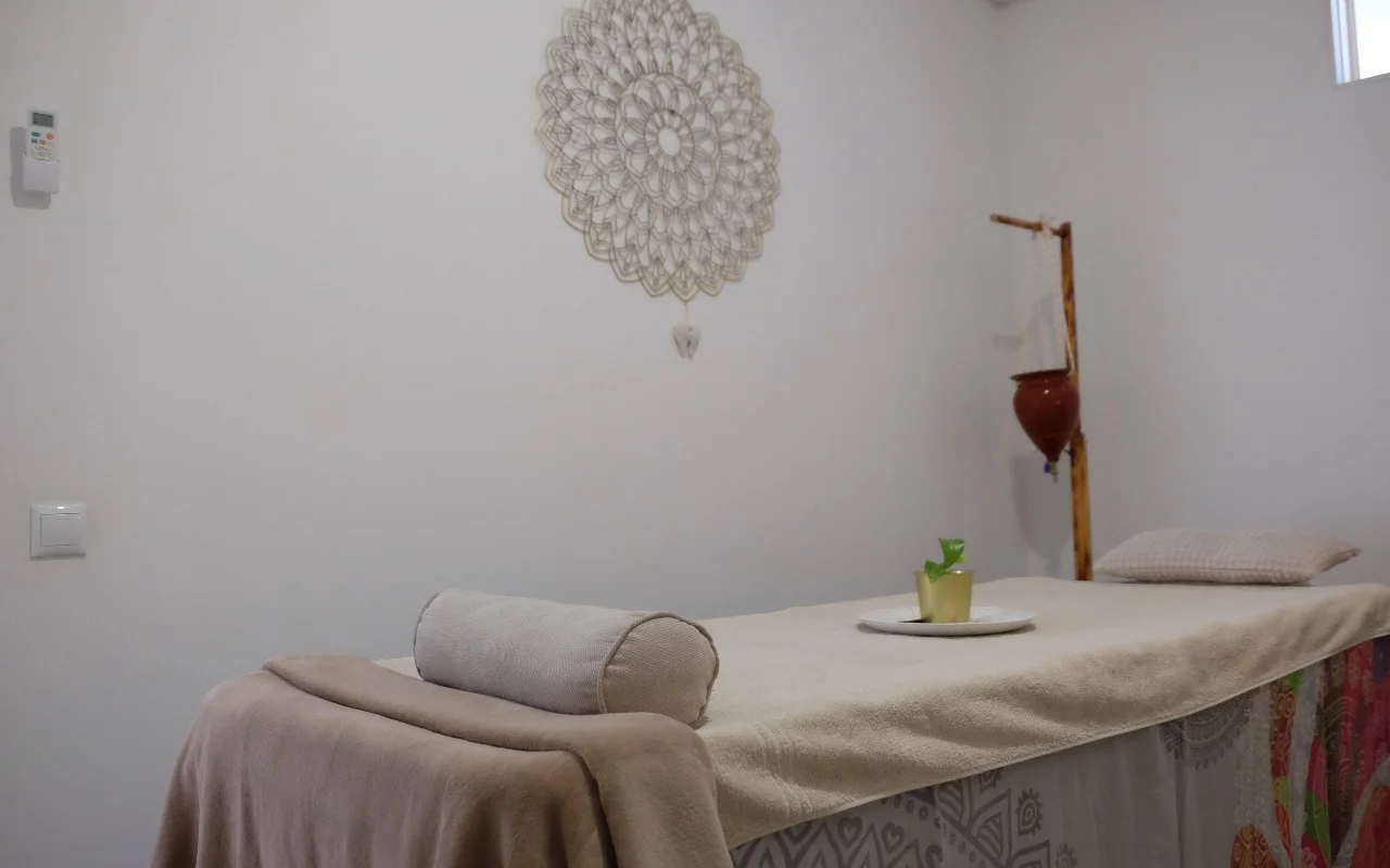 Una sala massaggi tranquilla in tonalità neutre e morbide, con un lettino da massaggio drappeggiato con un asciugamano e un cuscino a rullo, un grande mandala all'uncinetto appeso alla parete e un supporto in legno che regge un vaso di terracotta sospeso.