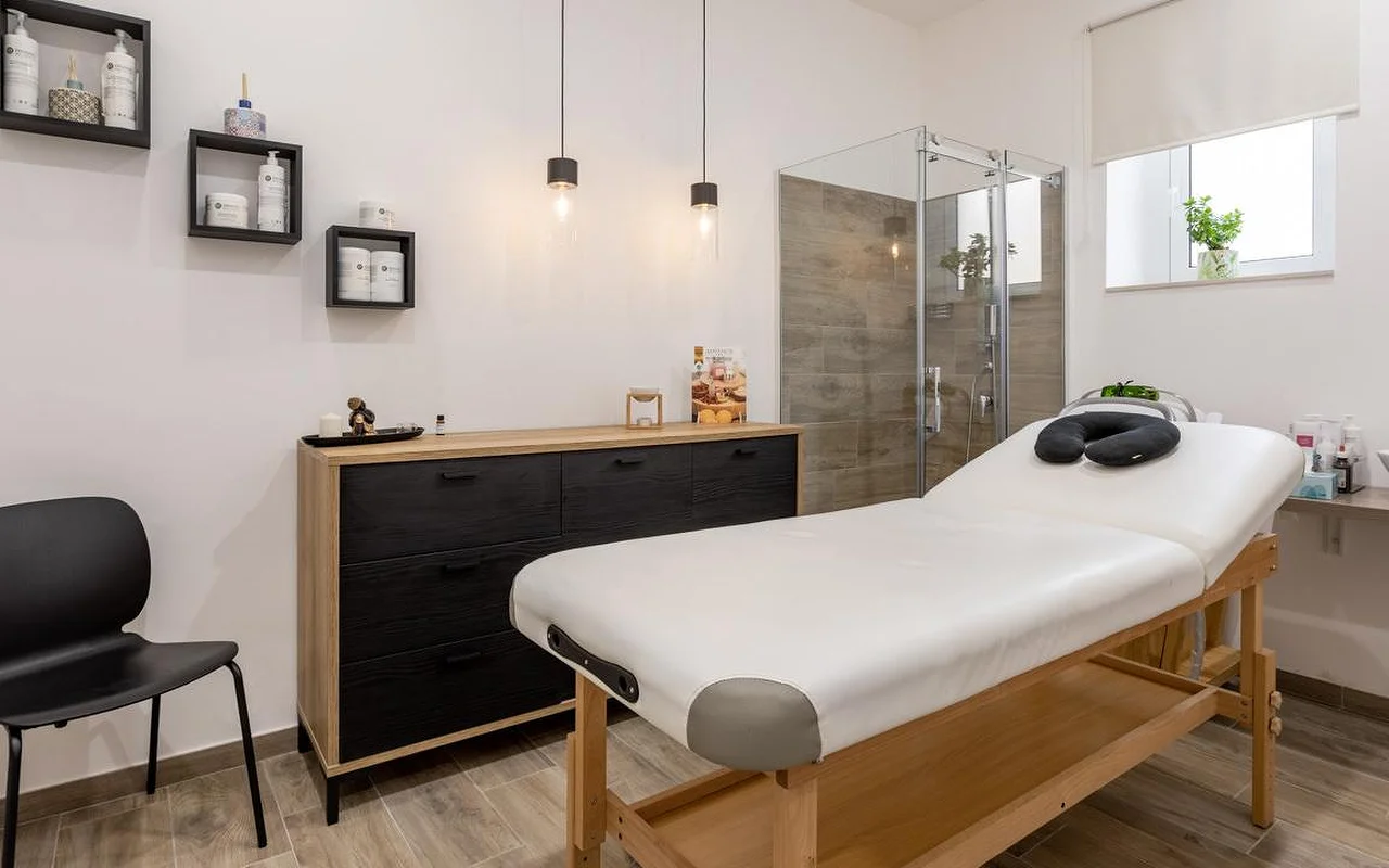 Una sala massaggi da Thea Estetica, con un lettino da massaggio in legno sormontato da un poggiatesta nero, un cassettone in nero e legno, due lampade a sospensione, mensole a cubo con i prodotti e un box doccia in vetro con piastrelle effetto legno.
