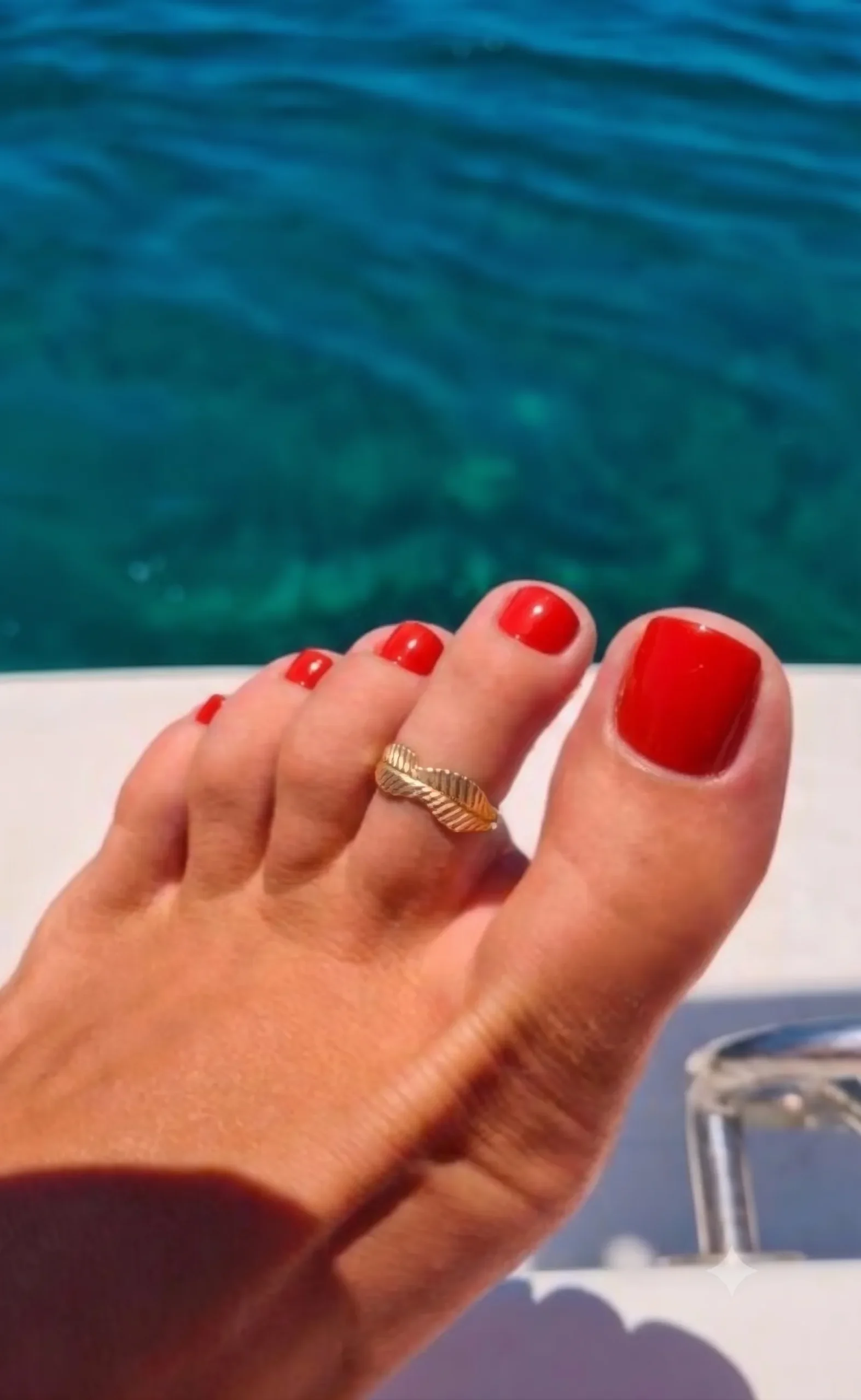 Pedicure rosso pomodoro lucida su un piede abbronzato con anello da piede dorato, mare turchese sullo sfondo