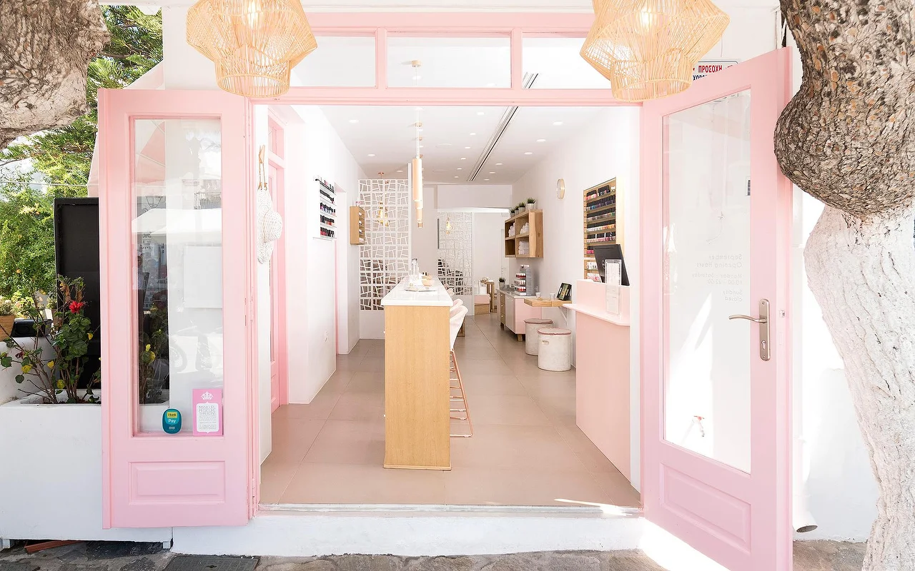 L'ingresso incorniciato di rosa di Vivian Nail Boutique a Naoussa, Paros, con porte aperte, lampade a sospensione intrecciate e bancone in legno, in un edificio cicladico imbiancato a calce accanto a un albero.