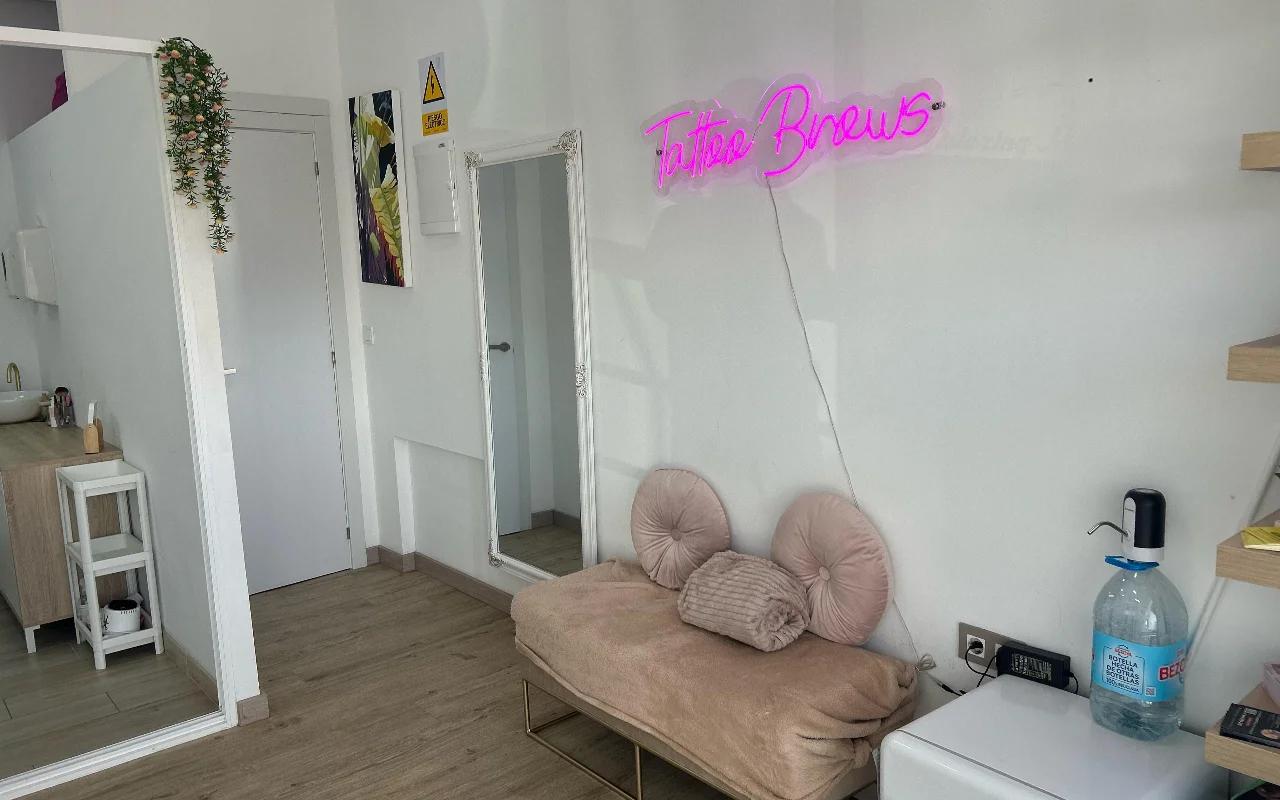 Uno spazio trattamenti da YG Beauty Estudio a Torremolinos, con un'insegna al neon rosa «Tattoo Brows», uno specchio a figura intera e un lettino beige imbottito.