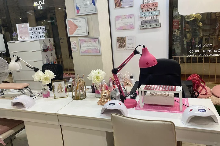 Postazione manicure di un salone unghie con scrivanie bianche, una lampada rosa acceso e lampade LED, rose bianche e certificati incorniciati alla parete, con il nome Color Nails PT visibile sulla vetrina accanto agli orari di apertura.