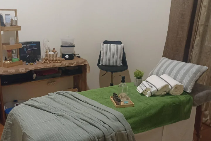 Cabina di massaggio accogliente con un lettino coperto da un asciugamano verde e da asciugamani bianchi arrotolati, un vassoio di oli essenziali, strumenti da massaggio in legno su un tavolino rustico, e uno sgabello accanto a un angolo separato da una tenda.