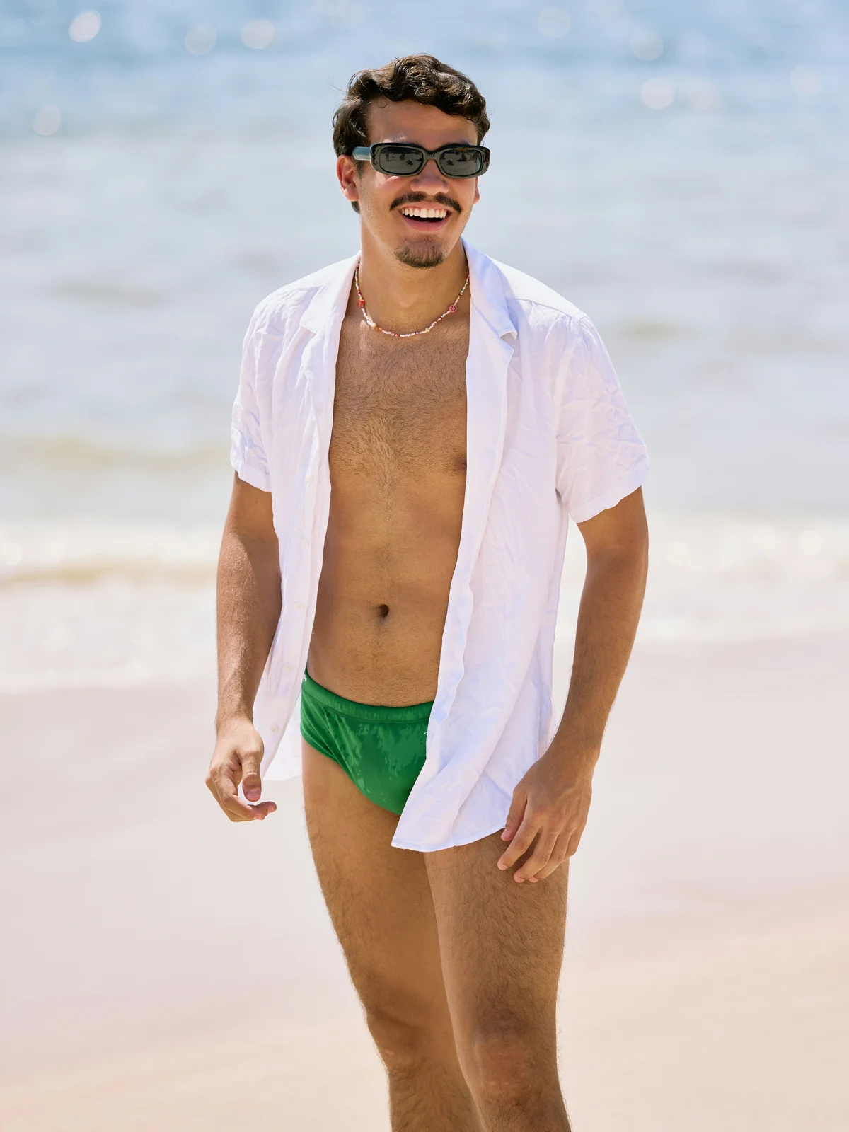 Un uomo sorridente è in piedi su una spiaggia in una giornata di sole; indossa un costume verde, una camicia bianca aperta e occhiali da sole neri