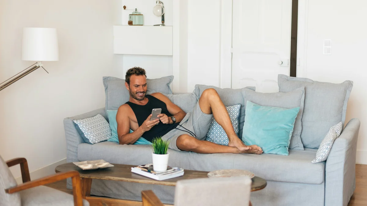 Un uomo si rilassa con i piedi sollevati sul divano e sorride mentre guarda il telefono; indossa una canotta nera e pantaloncini grigi morbidi e comodi