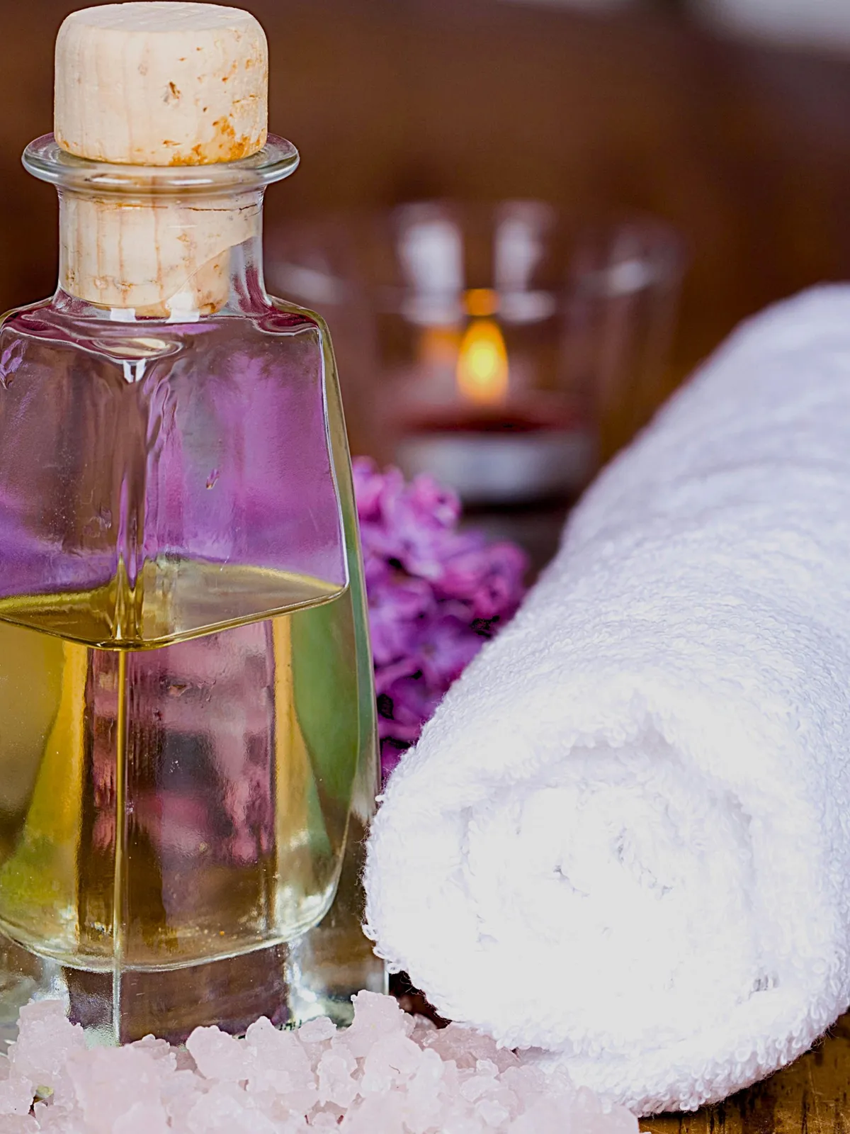 Primo piano di una bottiglietta di olio in vetro con tappo di sughero su un tavolo, accanto a un asciugamano bianco arrotolato e a fiori rosa
