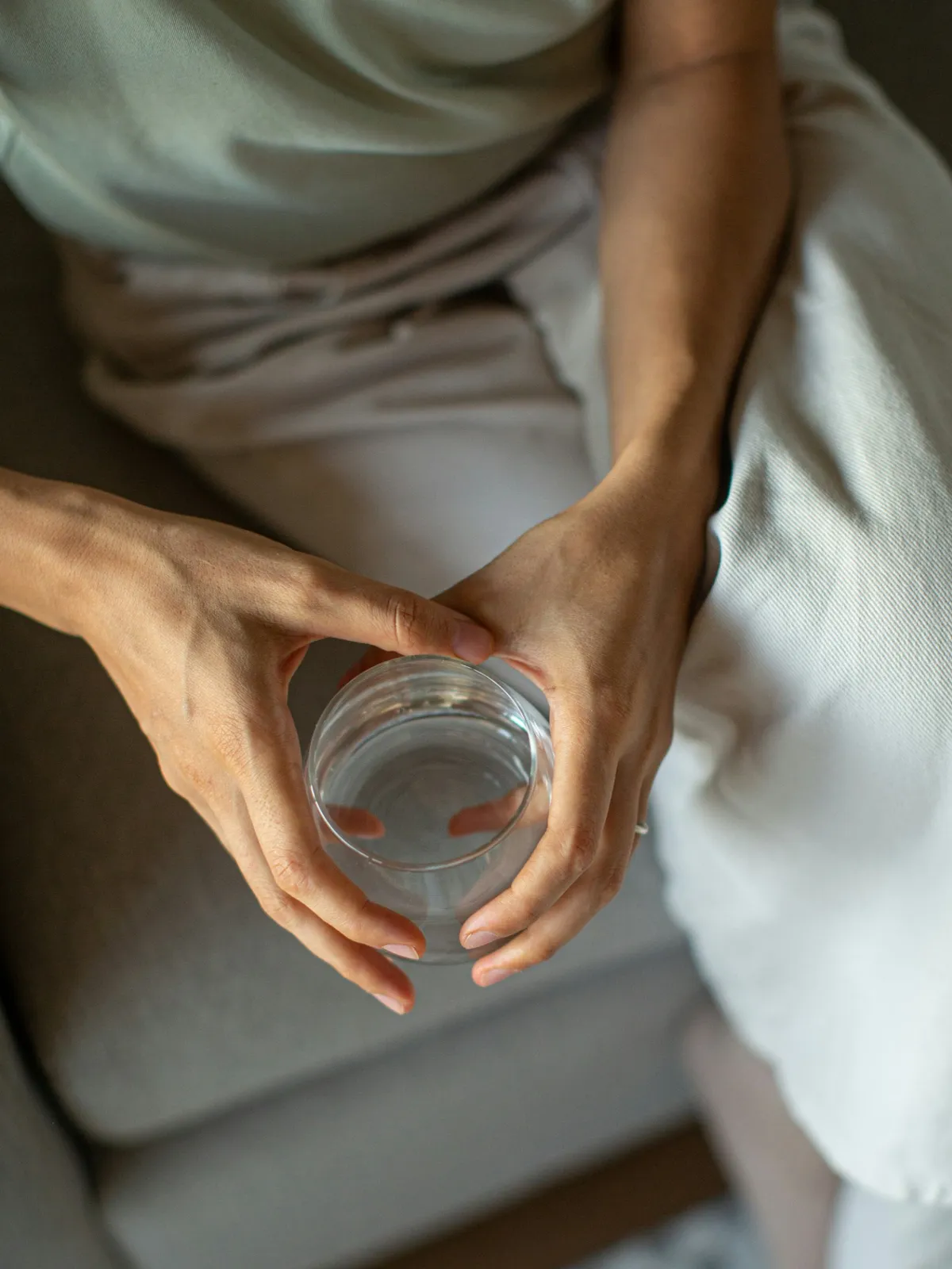 Veduta dall'alto di una persona in abiti comodi seduta su un divano grigio, che tiene un bicchiere d'acqua con entrambe le mani